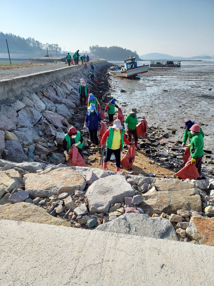 바다가 일대에서 초록색 옷과 모자를 쓴 사람들이 주황색 포대기 자루와  긴 집게를 들고 쓰레기를 줍고 있는 사진